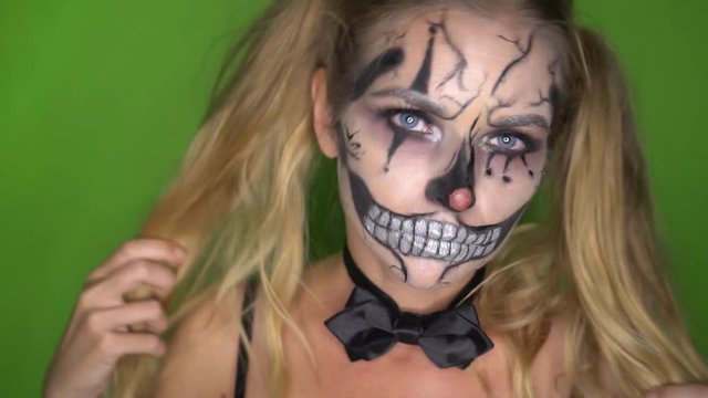 Closeup face of woman with creepy Halloween clown skull makeup looking into the camera standing over green screen background. Creative, artistic, Halloween concept - video in slow motion