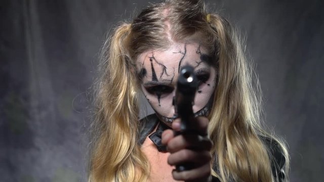 Closeup face of woman with creepy Halloween clown skull makeup looking and pointing the gun into the camera. Creative, artistic, Halloween concept - video in slow motion