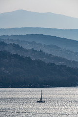 Obraz premium Barca in controluce al tramonto, con montagne a profili sfumati con luci cangianti, tra nebbie e freddo