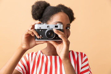 Stylish african american woman smiling and photographing on retro camera, isolated over beige background