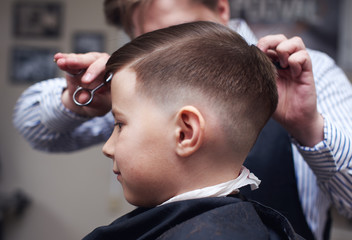 Barber making hairstyle to a Caucasian boy using scissors and hairbrush. - 228186294