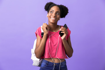 Photo of excited african american woman student wearing backpack and white headphones holding smartphone, isolated over violet background