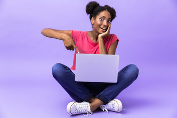 Photo of amusing african american woman smiling and using silver laptop, while sitting on floor with legs crossed, isolated over violet background