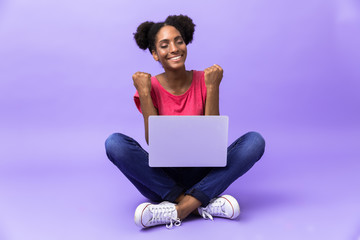 Photo of excited african american woman smiling and using silver laptop, while sitting on floor with legs crossed, isolated over violet background