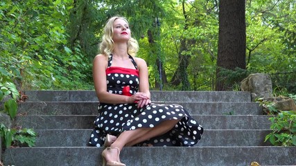 A young beautiful woman in a retro dress sits on a stony staircase in a park and looks around