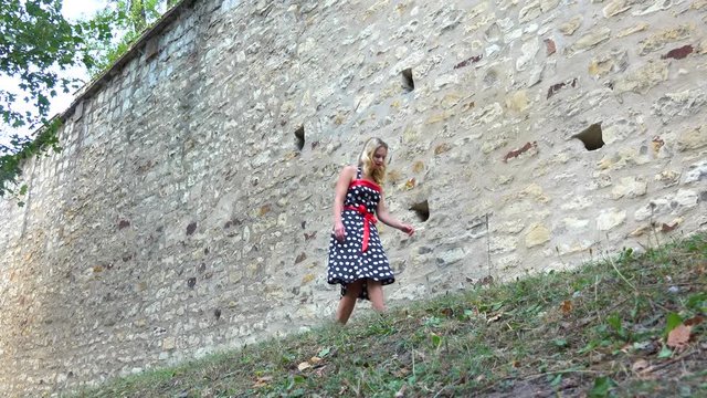 A young beautiful woman in a retro dress walks alongside a high, stone wall - view from below