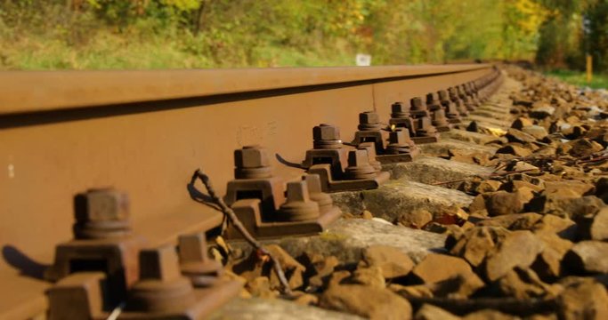 A close-up view of the large screws securing the train tracks during a sunny day with a slow passage forward with a turn from a side view from below a bit above the stones.
