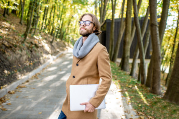 Portrait of a creative man in the coat and scarf standing with laptop outdoors in the forest during the autumn time