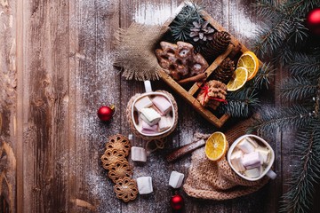 Christmas and New Year decor. Look from above at two cups with hot chocolate, cinamon cookies, green tree branches and snow on a wooden table