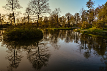 Fototapeta na wymiar Beautiful afternoon light in public park with green grass and trees reflecting in a pond. Shot in Cesis, Latvia.