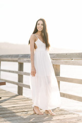 Fototapeta na wymiar Portrait of a smiling young woman in a white summer dress standing on a pier during the golden hour sunset.
