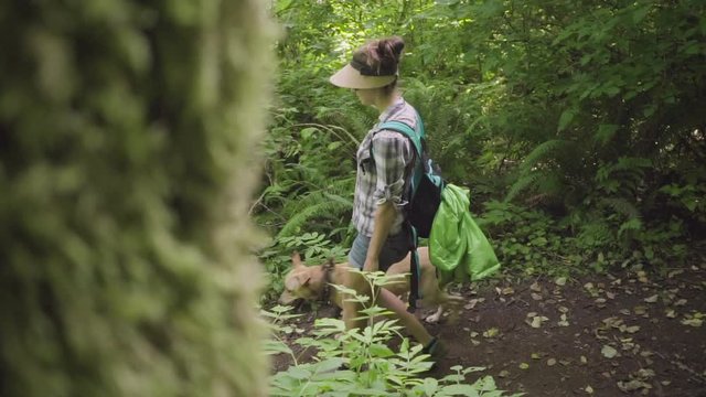 Young woman and dog walking down a hiking trail in a forest with a mossy tree in the foreground.