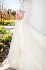 A bride holds up the train of her dress as she walks through a garden of flowers.