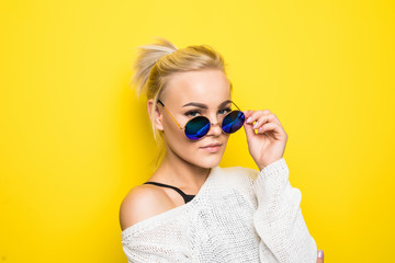 Smiling blond young woman in sunglasses posing and looking away on yellow background.