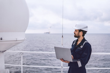 Marine service technician or serviceman near VSAT terminal on deck of vessel or ship. He is working on laptop or notebook