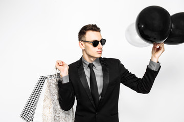 Fototapeta premium An elegant guy in a black suit and sunglasses, with shopping bags and balloons, is waiting for black Friday, on a white background. Black Friday