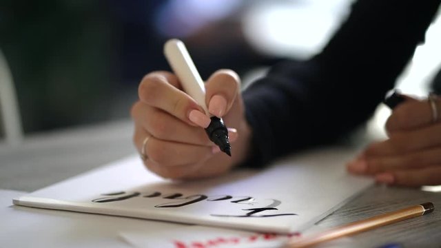 Calligrapher hands writes phrase on white paper. Inscribing ornamental decorated letters. Calligraphy, graphic design, lettering, handwriting, creation concept. Slow motion.