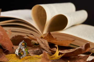Close-up Jewelry gold diamond ring on autumn foliage and open book pages in heart shape as background with copy space.Elegant engagement diamond ring as  love and wedding concept, close-up.