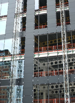 close up view of a modern construction site with hoists running up the tall building with steel girders with black panels and windows being installed
