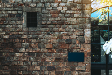 A stone wall of an ancient house made of masonry with a small window with bars above and empty blue nameplate template on the corner for the building number and a street name, Barcelona, Spain