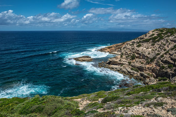 Fototapeta na wymiar Capo Mannu Cliffs in Sardinia, Italy against turquoise blue sea or ocean with blue sky and dramatic clouds. Waves crashing against rocks. 