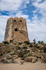 Fototapeta na wymiar Old watchtower on the beach of Torre di Bari, Sardinia, Italy. Beautiful blue sky with clouds in the background, a path visible and some unrecognizable people.