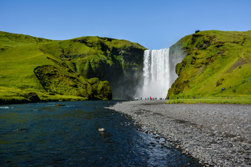 Famous Skogafoss Waterfall, Iceland with colourful tourists visible by water, sunny summer day, blue sky