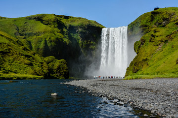 Fototapeta na wymiar Famous Skogafoss Waterfall, Iceland with colourful tourists visible by water, sunny summer day, blue sky