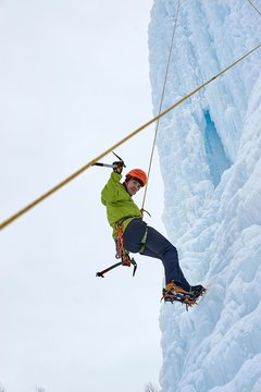Alpinist man with ice tools axe in orange helmet climbing a large wall of ice. Outdoor Sports Portrait.