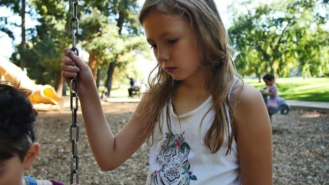 Blonde girl hanging out with friends on swing-set looks around playground as her friend is bullied