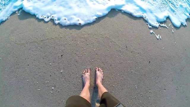 Male standing on a sandy beach and letting the waves roll up to his feet.