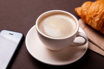 Coffee break, morning coffee. Cup of coffee with foam and croissant with smartphone.