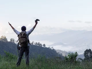 Tuinposter Happy male hiker with raised hands  and holding a binoculars standing on top of mountain © AungMyo