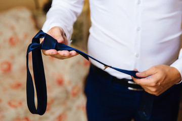 Close-up of a cropped frame of a slender business man in a trendy suit wearing dark trousers and a white shirt is holding a tie in his hands. A businessman is going to try on a blue tie.