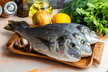 Three are ready to cook raw dorado fish on a wooden tray with herbs, lemon, onions, garlic, spices and olive oil on a gray textured background
