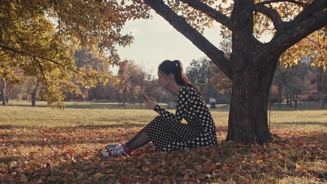 Female sitting under a tree in a prak using a phone.