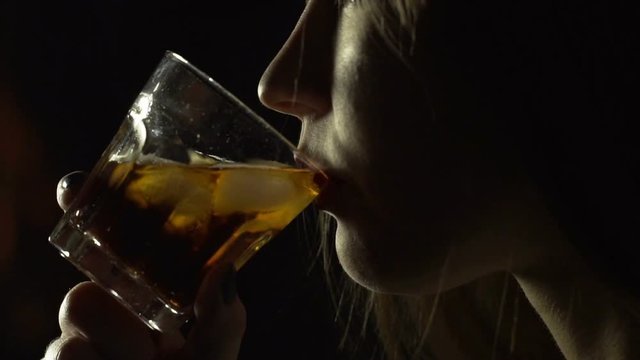 Girl drinks whiskey from glass on black background. Blonde girl tasting cognac