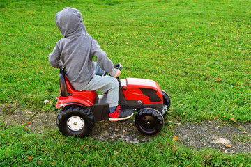 Little three years old boy driving red toy tractor outdoors in yard on green lawn grass background in autumn day back view. Children outdoor activities concept.