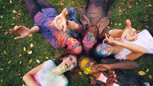 Top view of relaxed girls and guys relaxing on grass with painted faces after Holi festival looking at camera, smiling and moving hands. Recreation, fun and nature concept.