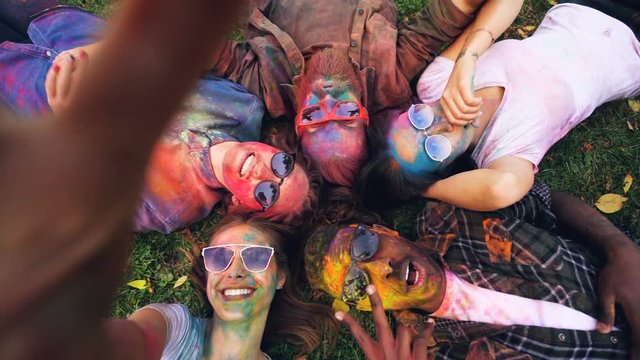 Slow motion point of view shot of beautiful young people with coloured faces lying on grass in circle looking at camera and posing with hand gestures.