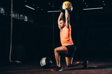 handsome fit sportsman performing lunge with medicine ball overhead in dark gym - obrazy, fototapety, plakaty