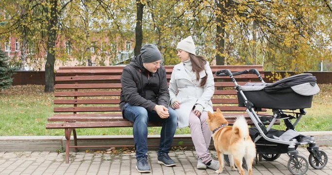 Happy family walking in autumn park with stroller. Mom, Dad and their newborn baby son sit on a bench and play with their family dog