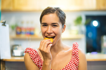 Portrait of happy young woman eating cookie in kitchen. Latin American woman biting pastry with...