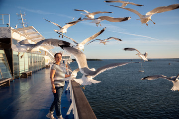 Older woman watching seagulls flying. Photo of a middle aged lady on the cruise ship deck in a Baltic Sea cruise. - obrazy, fototapety, plakaty