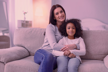 Warm relations. Joyful positive woman hugging her beloved daughter while sitting on the sofa