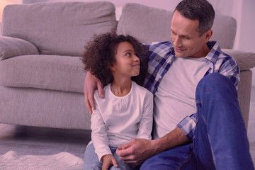 Happy fatherhood. Positive nice man hugging his daughter while sitting with her in front of the sofa