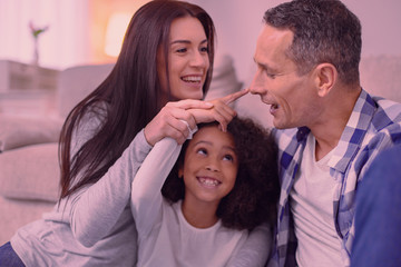 What is this. Joyful positive girl touching her dads nose with her finger while sitting between her parents