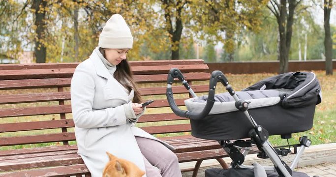 Young mother walking with her little baby son in stroller in autumn park and using mobile phone while her newborn baby sleep.