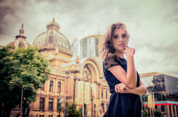 Fototapeta premium Beautiful blonde girl with big blue eyes and black dress and messed up hair, looking pensive, in the city