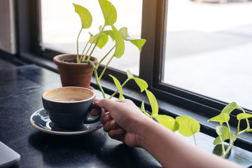 Closeup image of a woman's hand holding a cup of coffee with small tree pot on wooden table